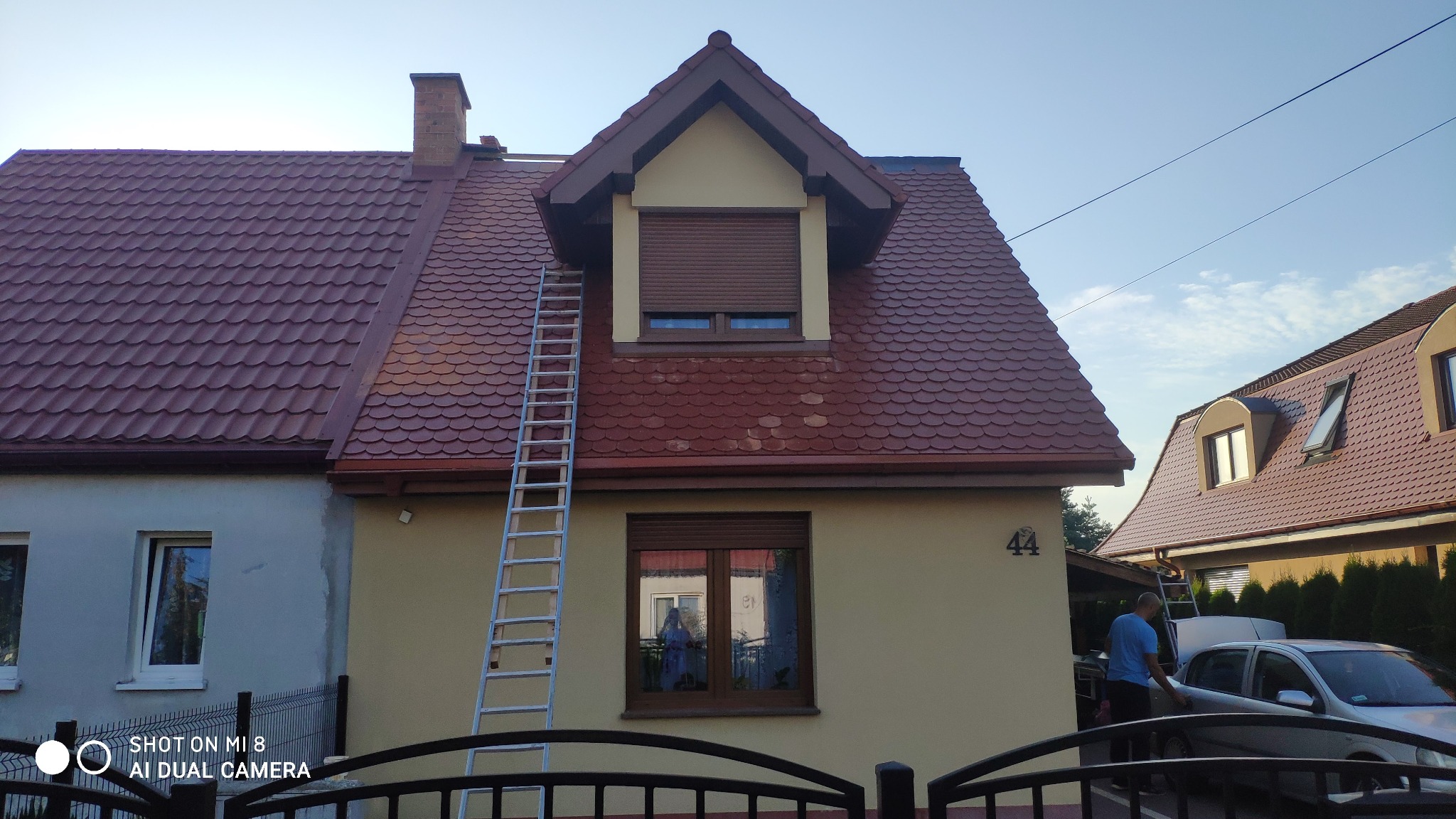 Dom jednorodzinny z nowym, bordowym dachem z blachodachówki, drabina oparta o okno na poddaszu, widoczne obróbki blacharskie, elewacja w kolorze kremowym.