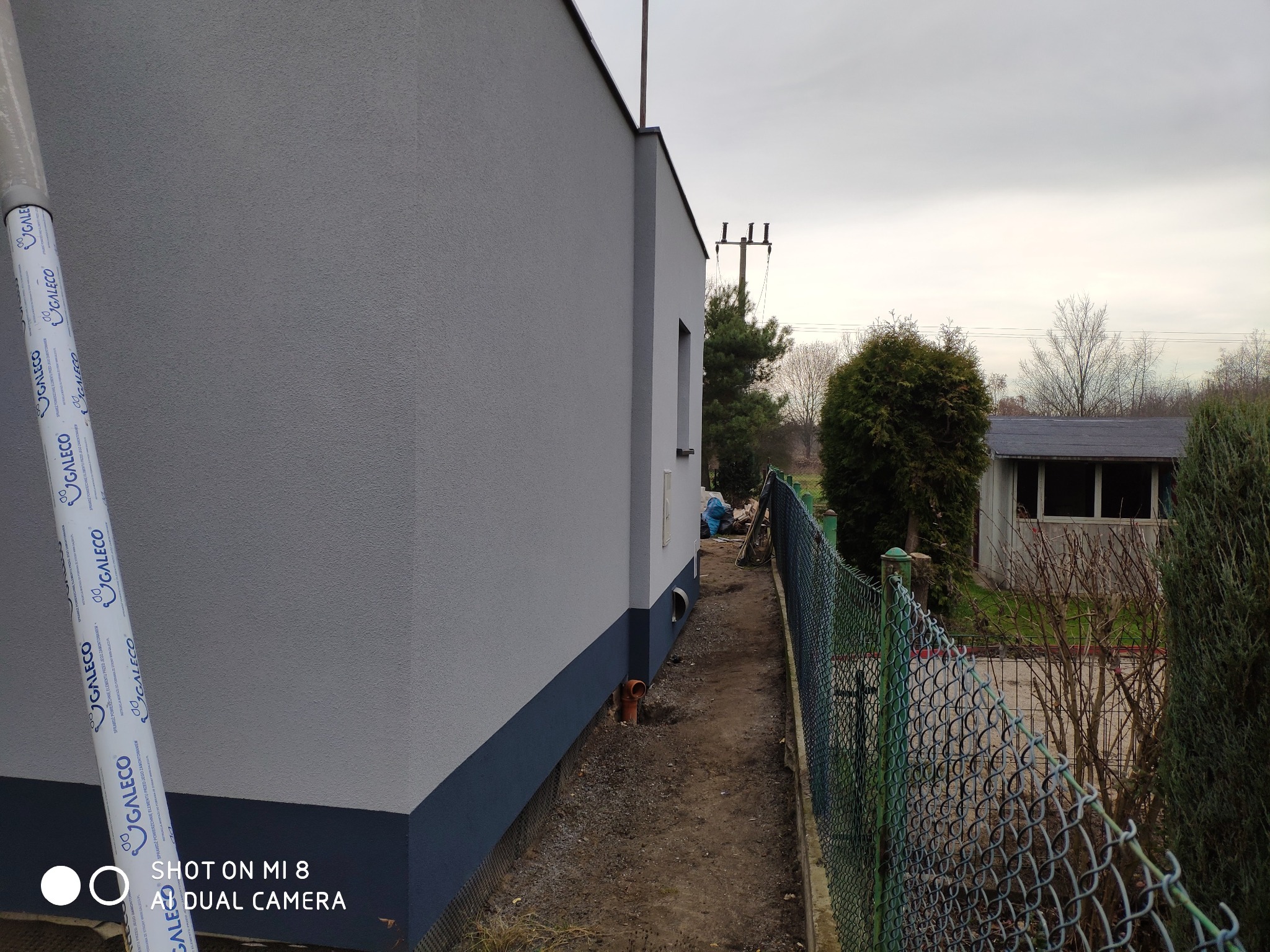 Świeżo otynkowana ściana domu w odcieniach szarości z widocznym fragmentem rolki folii ochronnej GaleCo i fragmentem rynny, zarys ogrodzenia z siatki, budynek gospodarczy w tle.