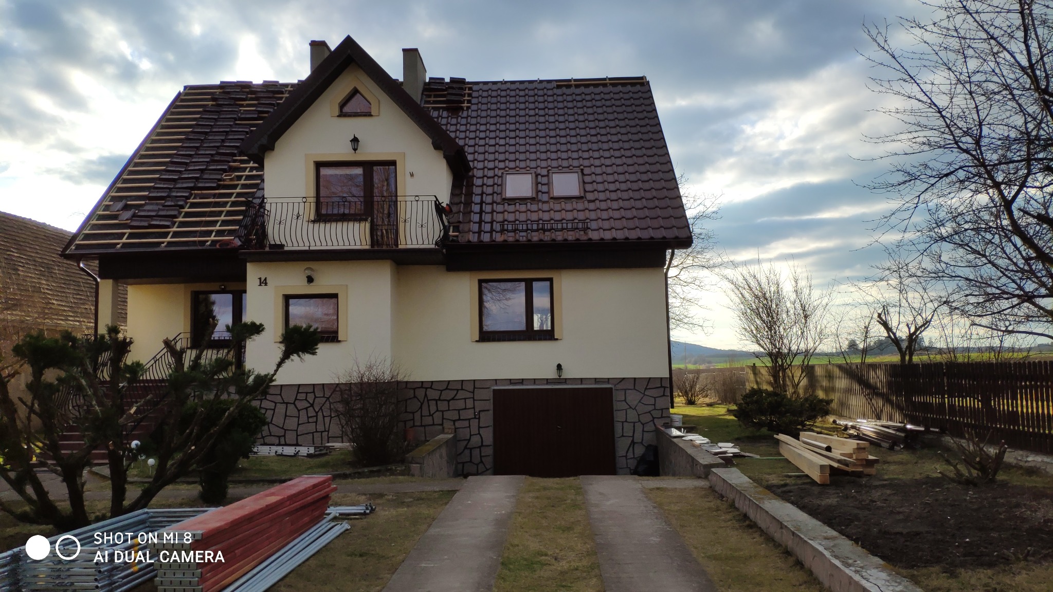 Dom jednorodzinny z częściowo zdjętym dachem, widoczne krokwie i nowa dachówka, w tle pochmurne niebo i drzewa bez liści.