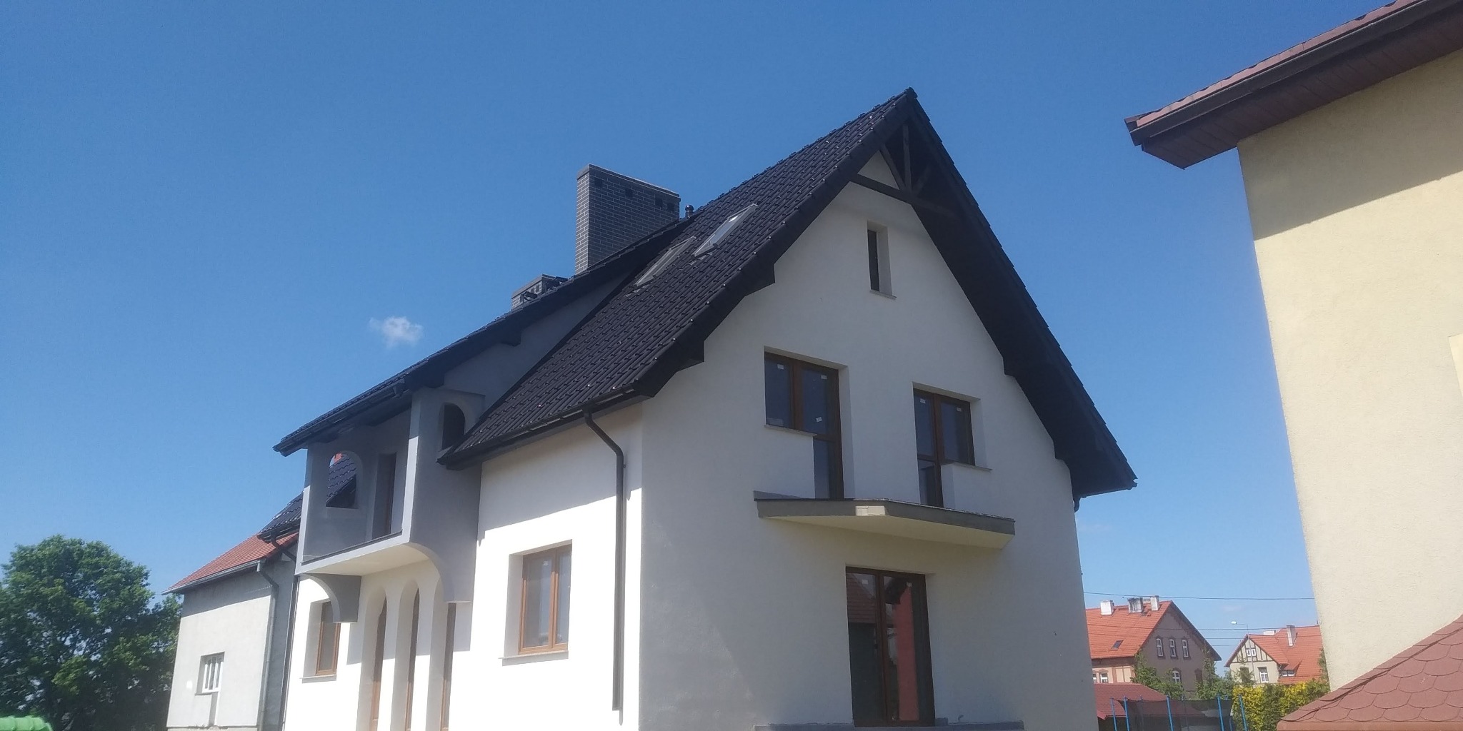 Nowoczesny dom jednorodzinny z ciemną dachówką i jasną elewacją, widok z dołu na tle błękitnego nieba.