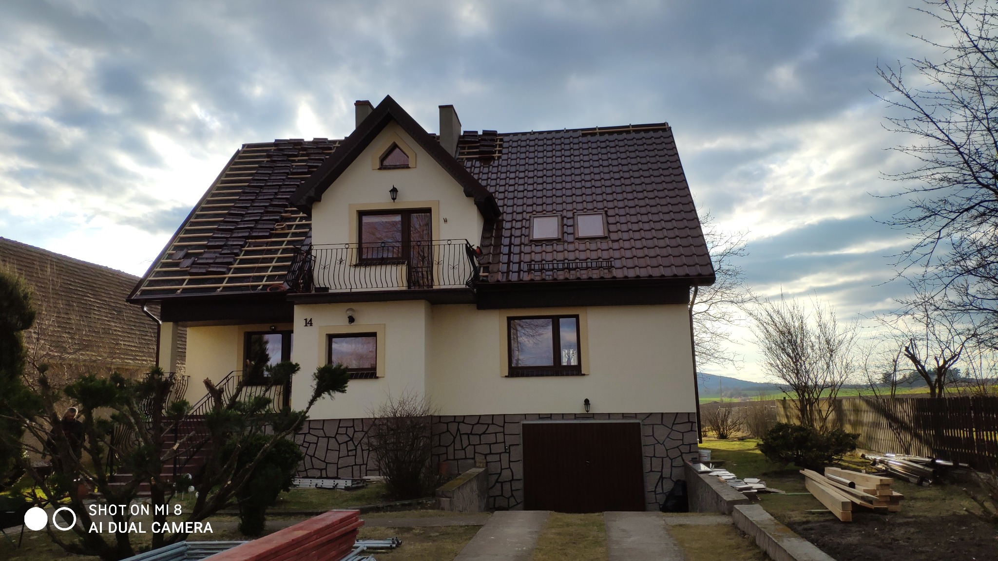 Dwukondygnacyjny dom z częściowo wymienionym brązowym dachem, widoczne łaty i nowe dachówki, elewacja w odcieniach beżu z kamienną okładziną na dole, balkon z ozdobną balustradą, pochmurne niebo...