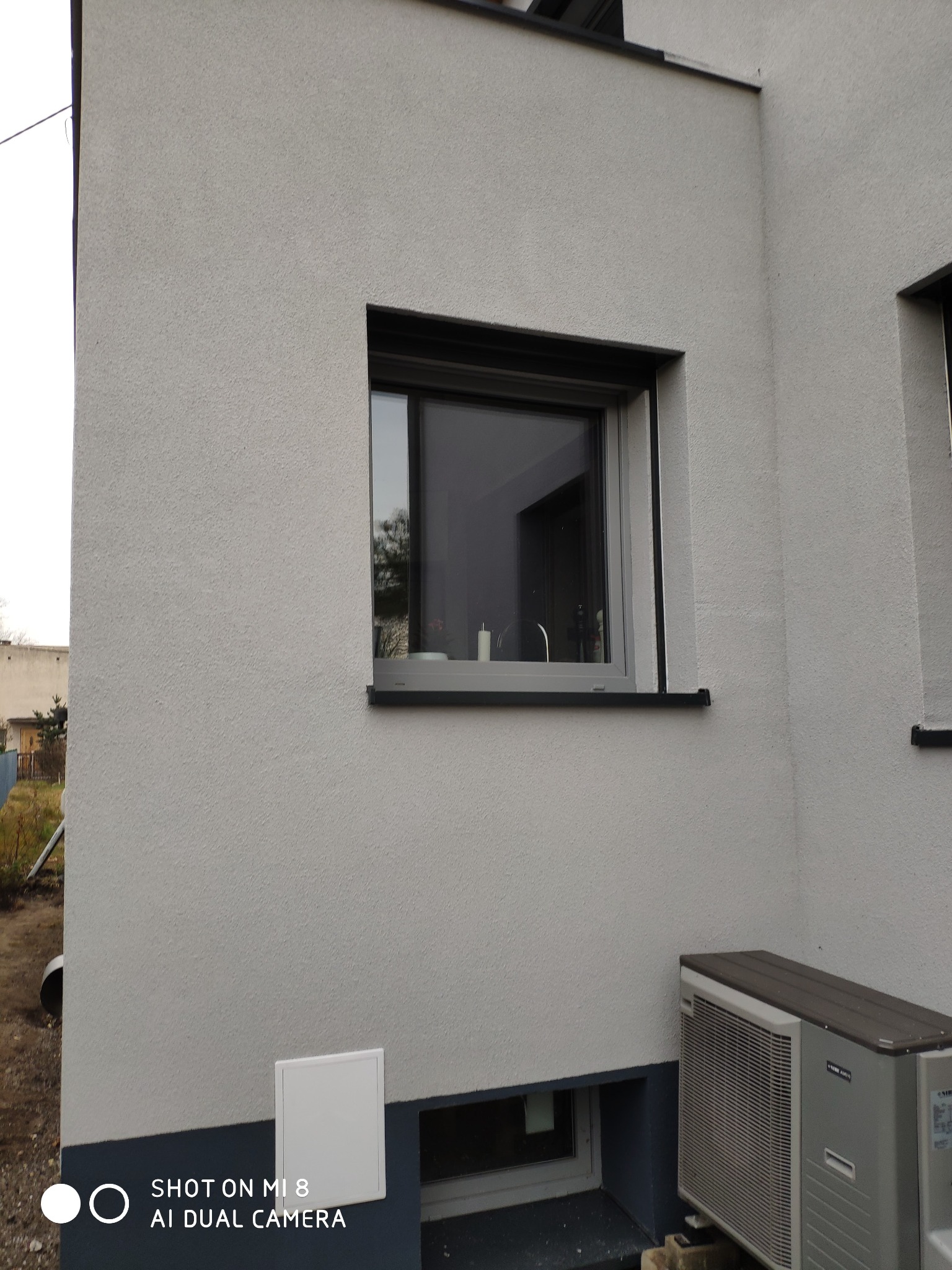 Szara elewacja budynku jednorodzinnego z oknem w czarnej ramie i zamontowanym klimatyzatorem zewnętrznym. Widoczny fragment cokółu budynku. Na elewacji widoczna faktura tynku.