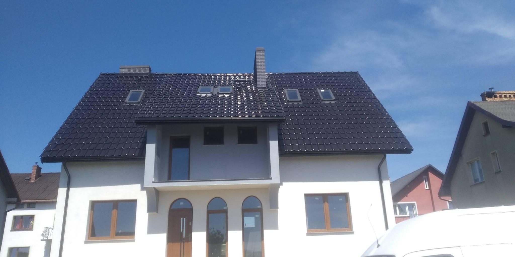 Dom jednorodzinny z nowym, błyszczącym dachem z dachówki ceramicznej w kolorze grafitowym, widoczne okna dachowe i komin z cegły, elewacja w kolorze białym, błękitne niebo w tle.