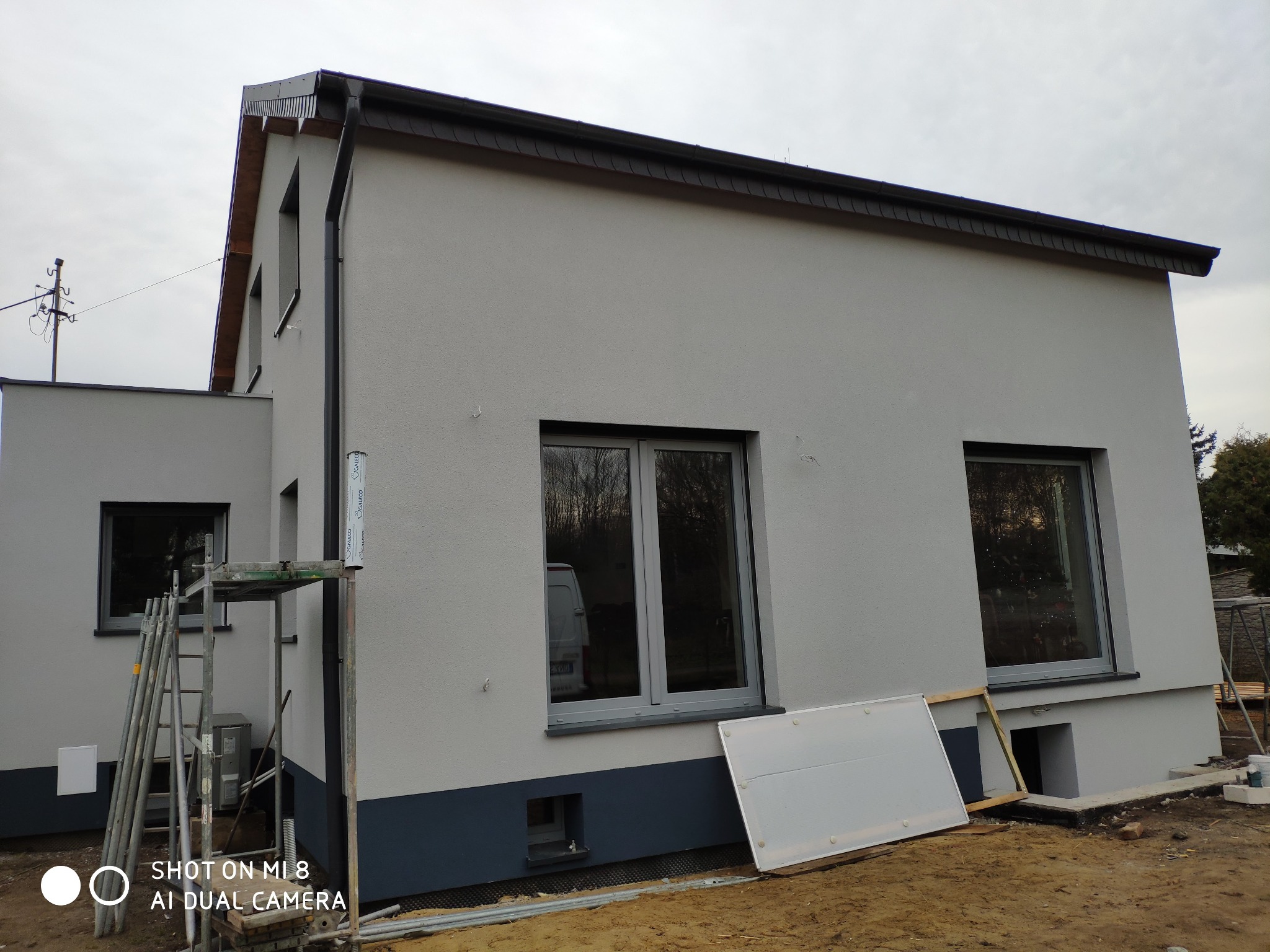 Nowoczesny dom jednorodzinny z szarą elewacją i grafitową podmurówką, widoczne rusztowanie i elementy przygotowane do prac wykończeniowych wokół budynku.