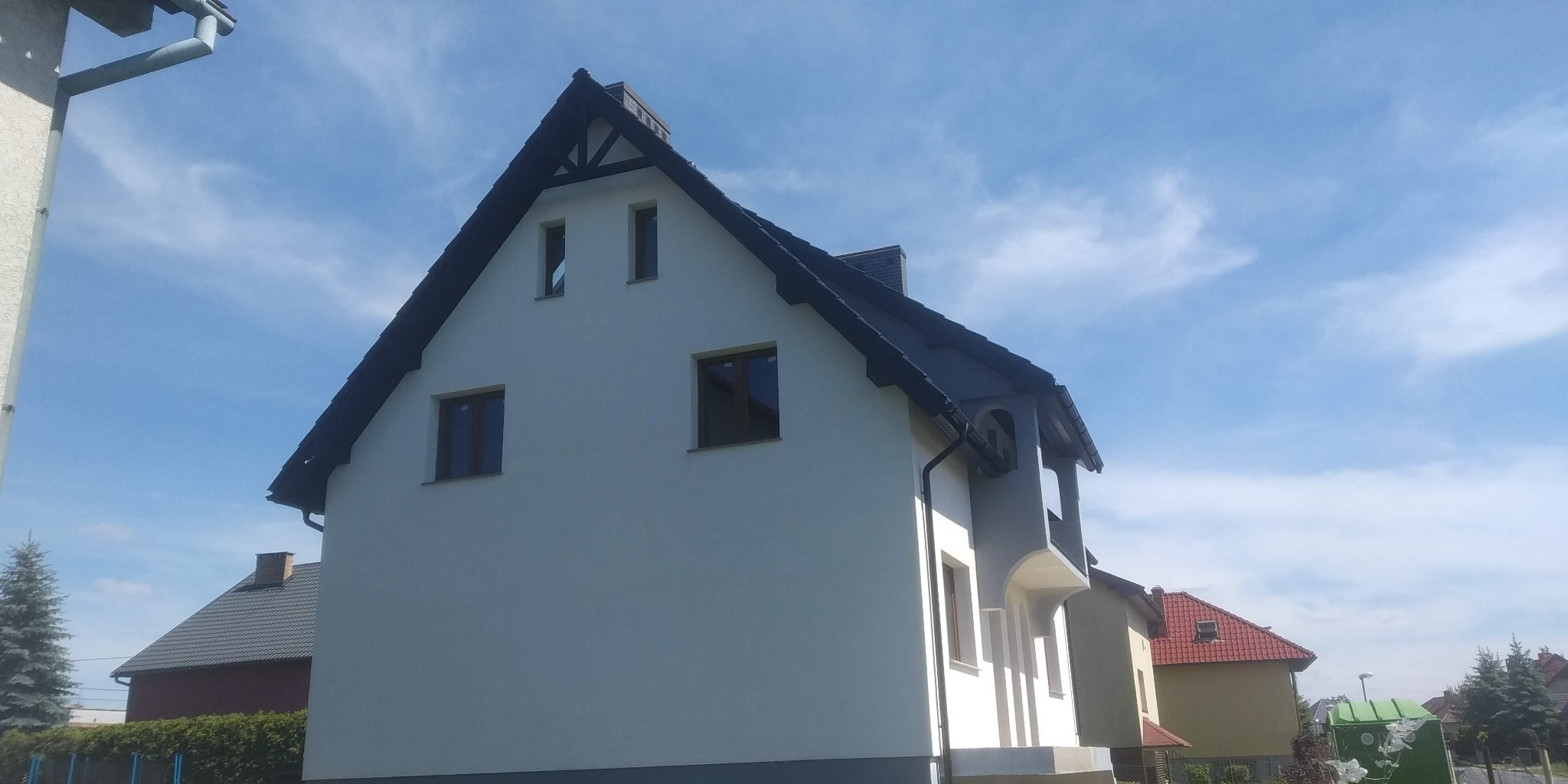 Dwukondygnacyjny dom z ciemnym dachem i jasną elewacją, widoczny z boku pod kątem, z fragmentem nieba w tle, z wyraźnie widocznymi oknami i elementami wykończeniowymi.