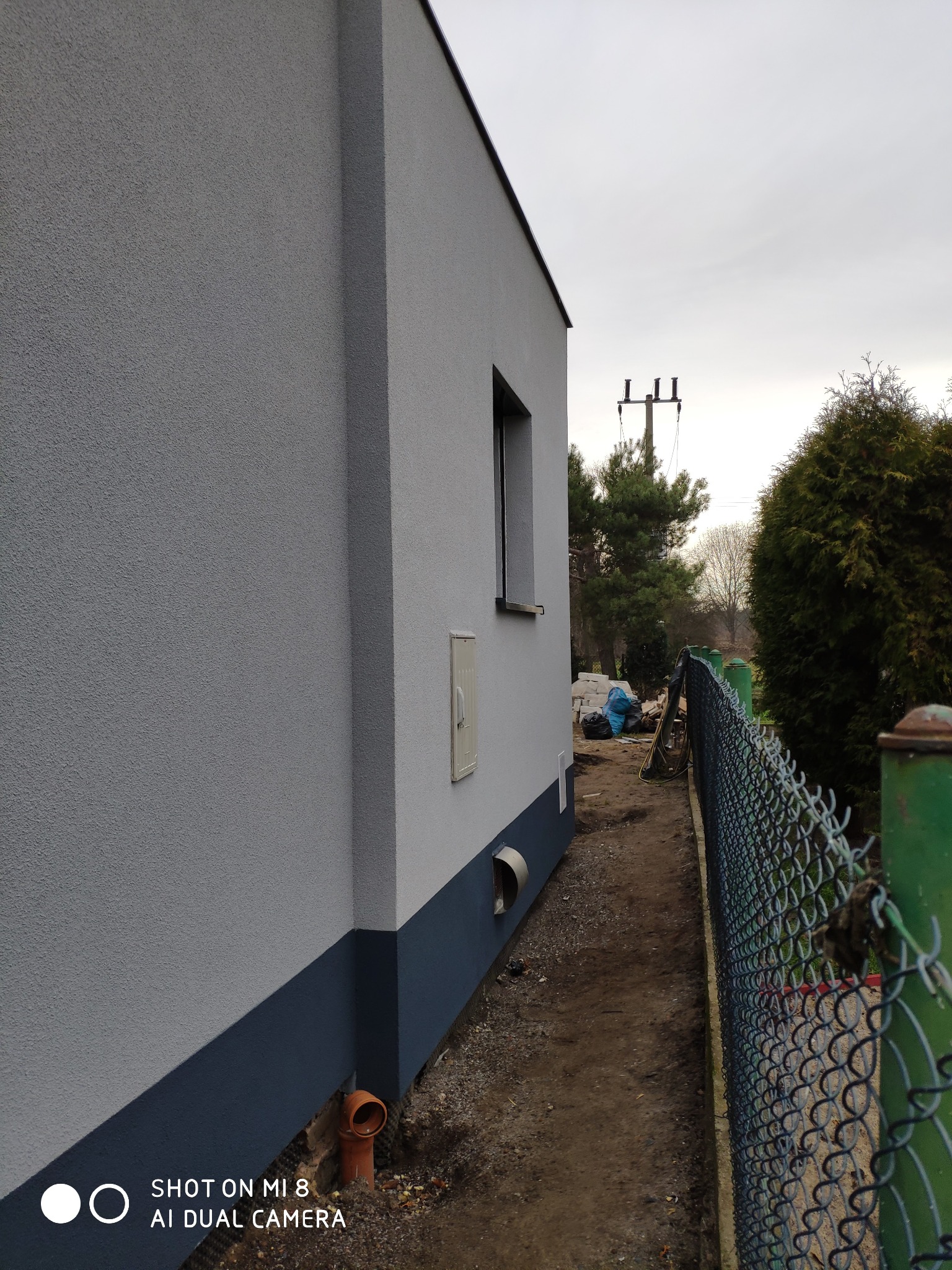 Świeżo otynkowana ściana budynku z widoczną podmurówką w kolorze ciemnego granatu, rura spustowa i fragment okna; w tle drzewa i słup energetyczny.