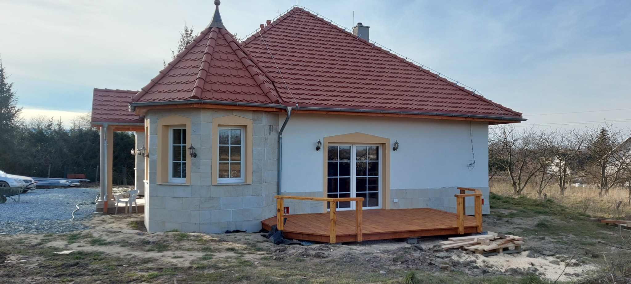 Nowoczesny dom z czerwonym dachem z dachówki karpiówki i drewnianym tarasem, elewacja w trakcie wykończenia.