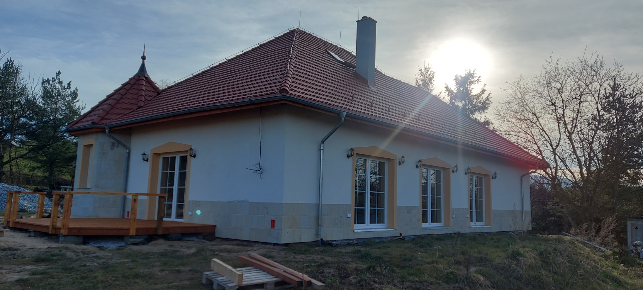 Nowoczesny dom jednorodzinny z nową, czerwoną dachówką ceramiczną, świeżo wykończoną elewacją w kolorze kremowym z dekoracyjnym kamieniem na fundamencie i drewnianym tarasem w trakcie budowy.