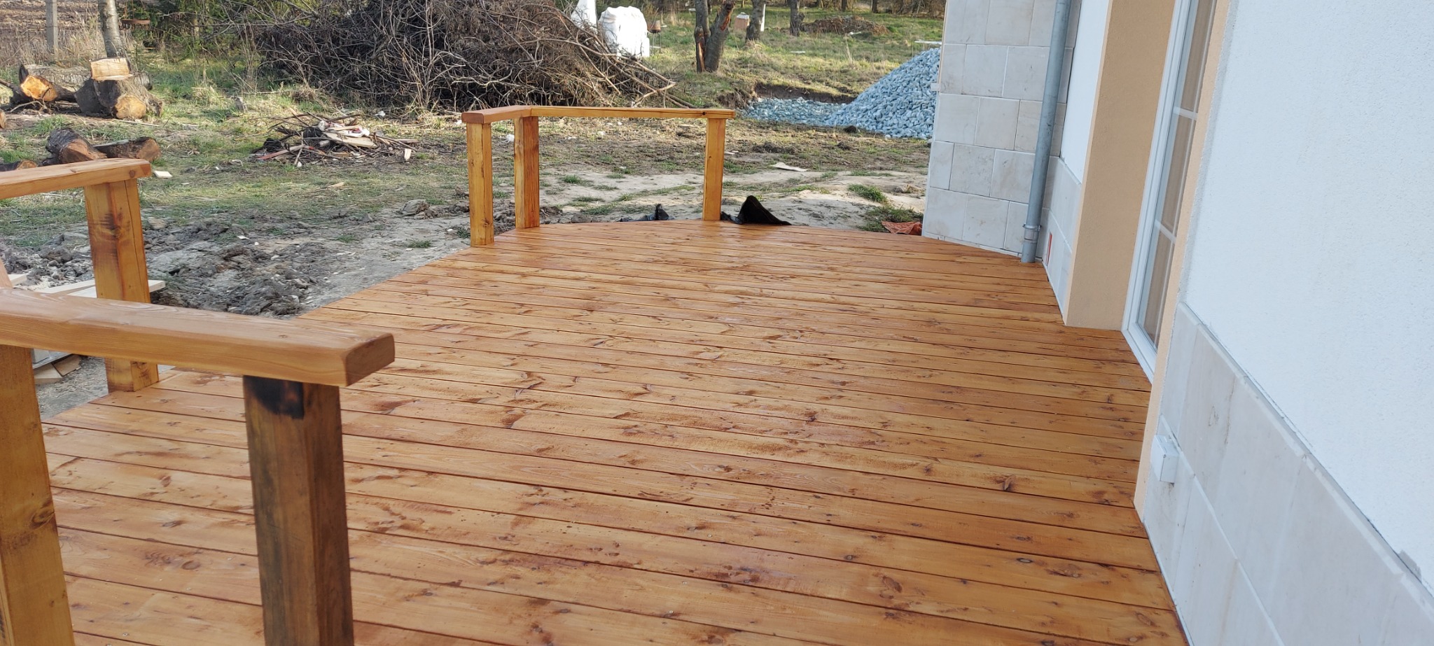 Nowa drewniana podłoga tarasu z balustradą, świeżo po impregnacji, przylegająca do ściany budynku z oknem. W tle widoczny stos drewna i kamieni.