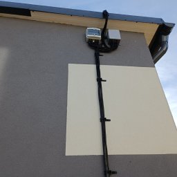 Energysat - Zewnętrzna instalacja elektryczna z licznikiem i skrzynką przyłączeniową, przymocowana do ściany budynku z szaro-białą elewacją i rynnami.
