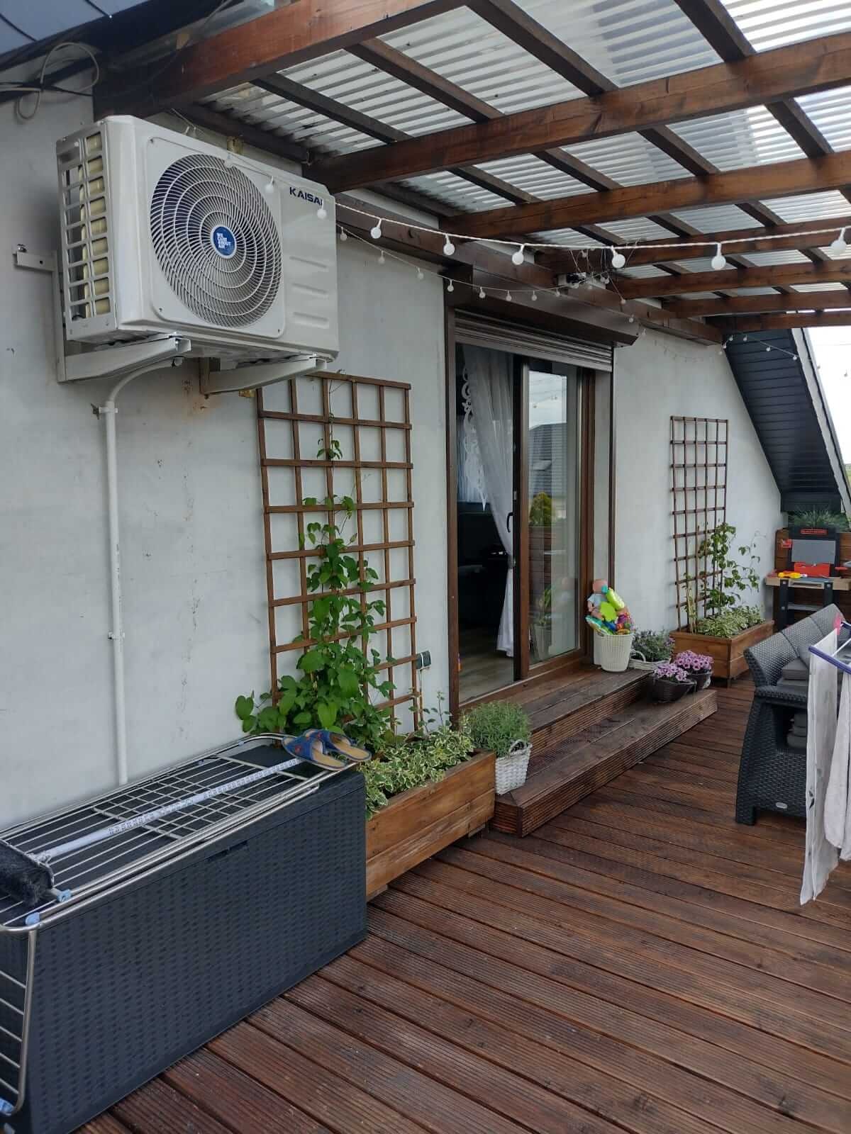 Biała jednostka zewnętrzna klimatyzacji marki Kaisai zamontowana na ścianie tarasu z drewnianą podłogą i zadaszeniem, obok kraty z pnącą rośliną i skrzynki z kwiatami.