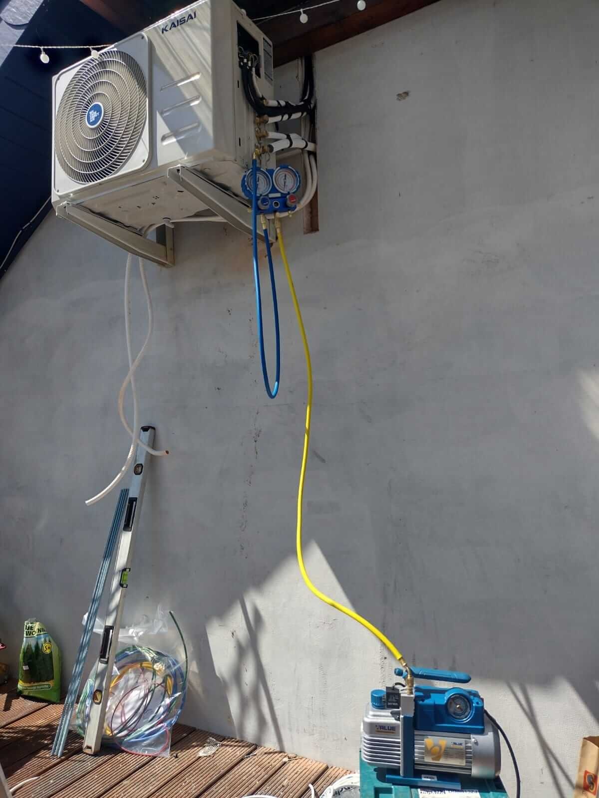 Biała jednostka zewnętrzna klimatyzacji Kaisai zamontowana na ścianie, podłączona do manometrów i pompy próżniowej za pomocą żółtego i niebieskiego węża, obok poziomica i zwój kabli w folii.