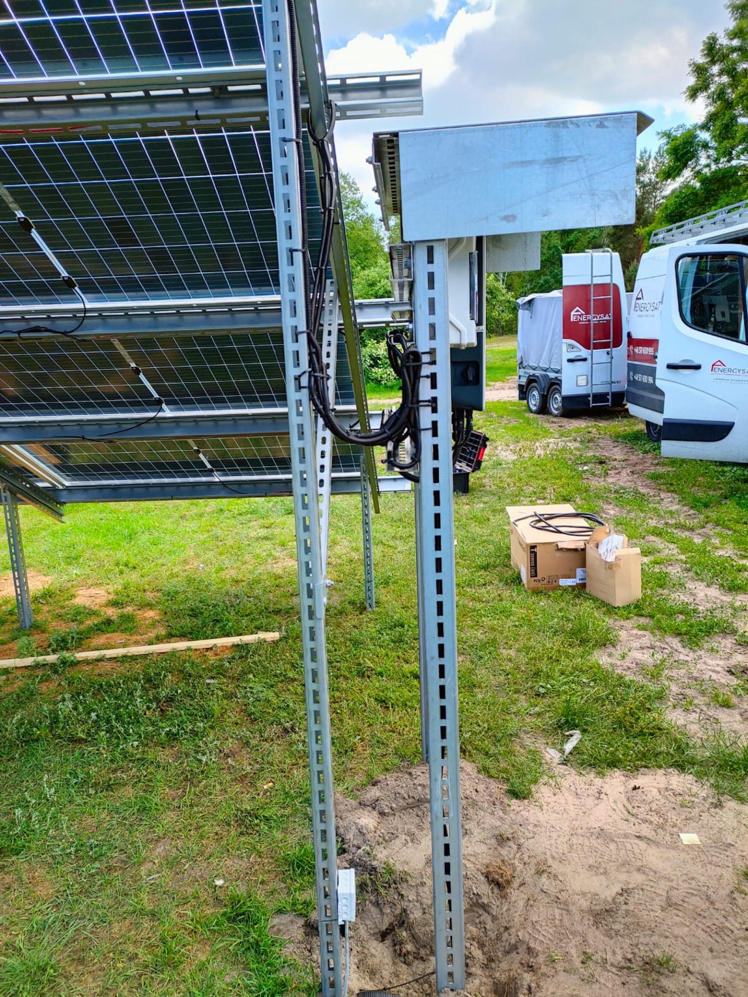 Montaż konstrukcji wsporczej paneli słonecznych na trawiastym terenie, widoczne metalowe profile, okablowanie, kartony po elementach instalacji i samochód dostawczy z logo firmy w tle.