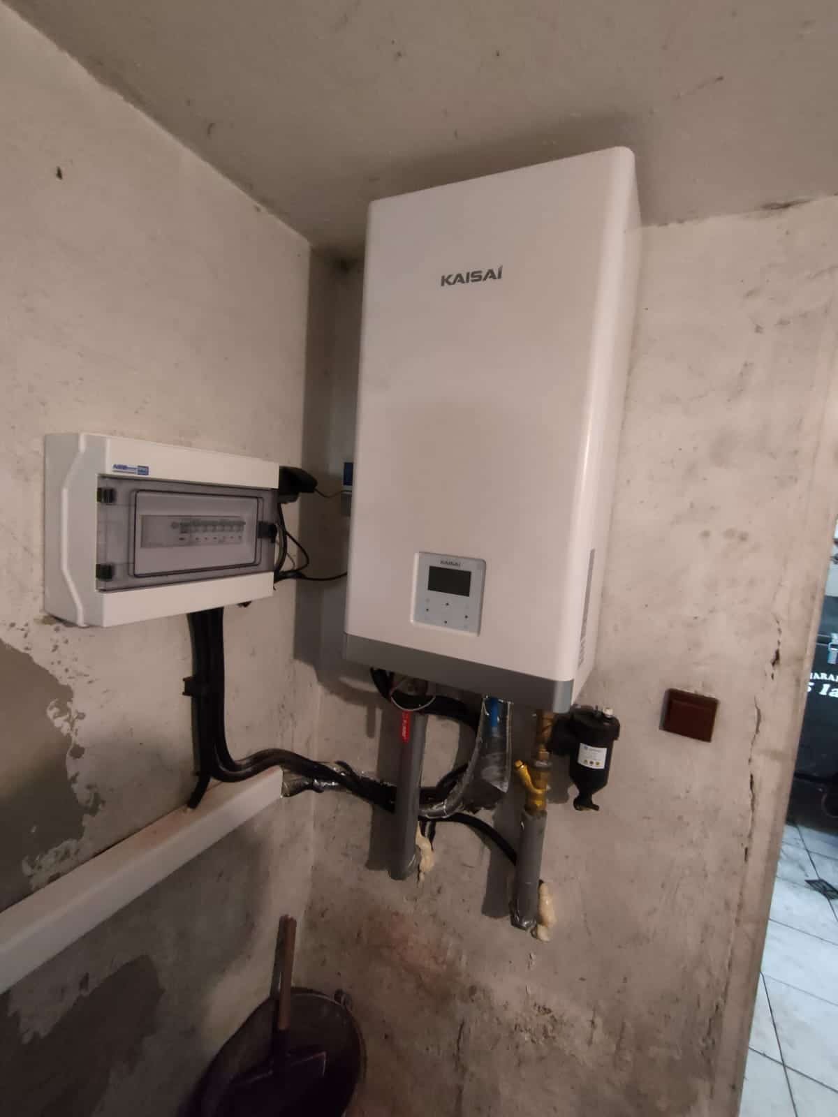 Biała jednostka wewnętrzna pompy ciepła marki Kaisai zamontowana na szarej, surowej ścianie piwnicy, obok skrzynki elektrycznej z widocznymi przewodami i rurami instalacyjnymi.