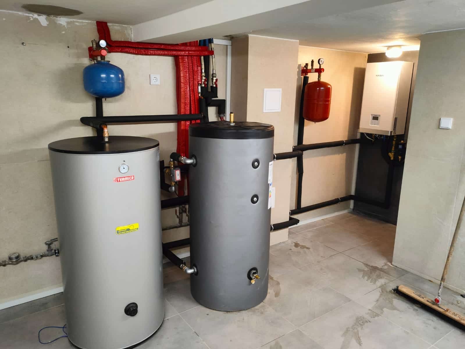 Dwa zbiorniki akumulacyjne ciepła (jeden szary, drugi srebrny z logo Termica) połączone rurami, z widocznym bojlerem i instalacją grzewczą w tle, na podłodze widoczne ślady wilgoci.
