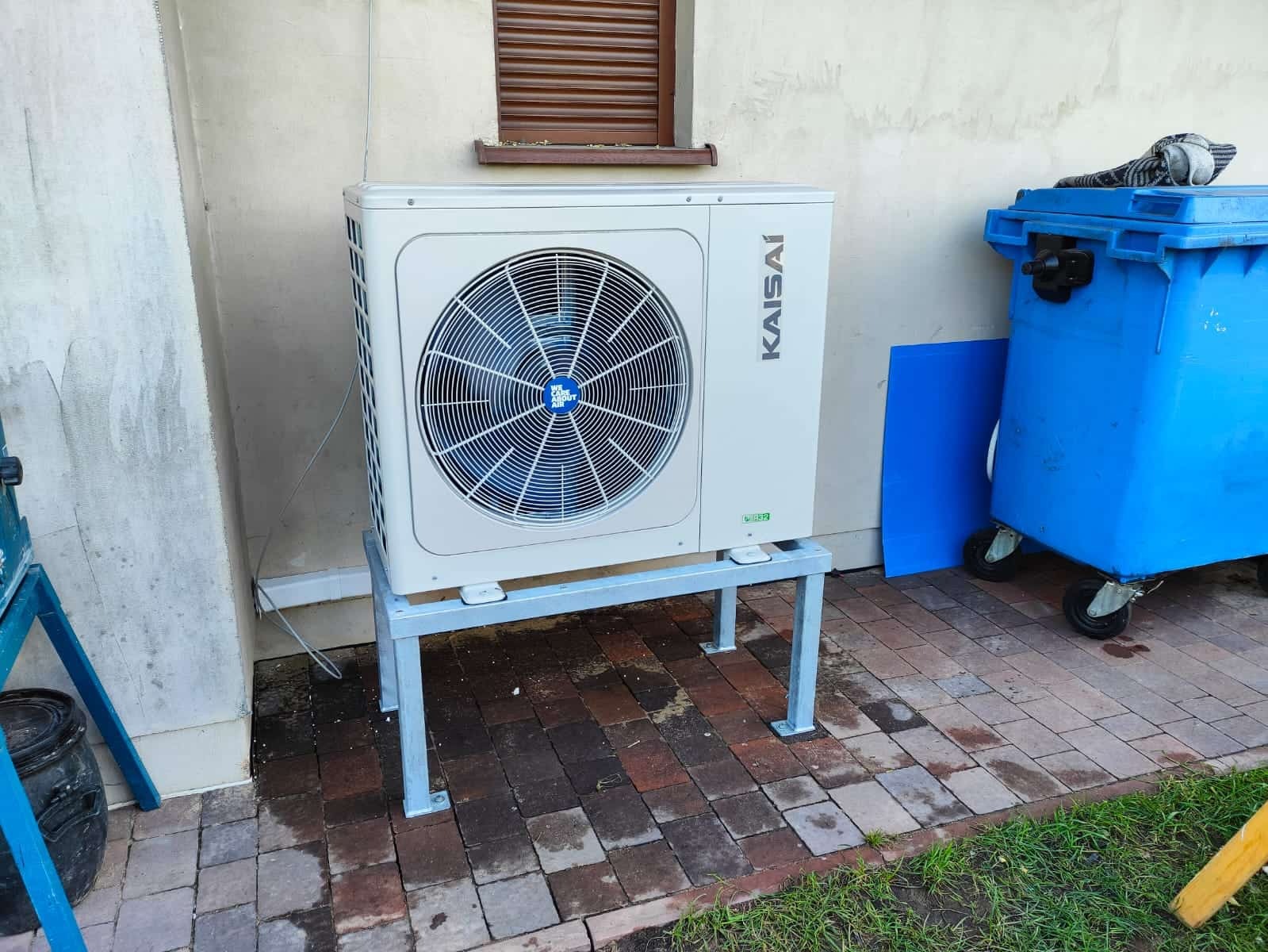 Zewnętrzna jednostka klimatyzacji Kaisai na metalowym stelażu, ustawiona przy ścianie budynku na kostce brukowej, obok niebieski pojemnik na śmieci z kołami.