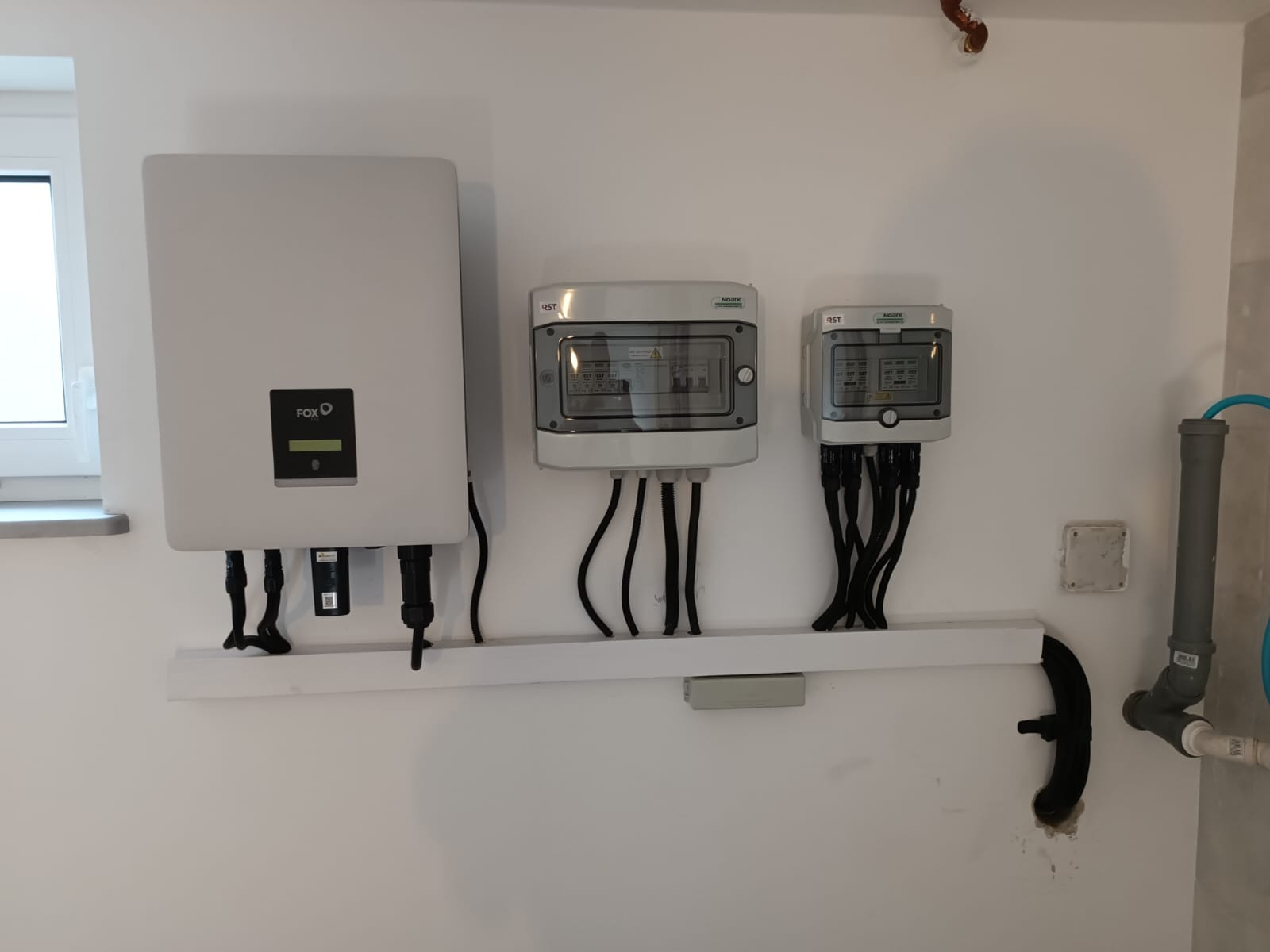 Biały inwerter FOX z czarnymi przewodami, dwie szare skrzynki rozdzielcze z przezroczystymi okienkami, przymocowane do białej ściany na białej listwie, obok okno i rury instalacyjne.
