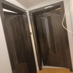 Dwa ciemne, lakierowane drzwi wewnętrzne z szarobrązowym fornirem, zamontowane w białej ścianie nad parkietem ułożonym w jodełkę, oświetlone centralnym plafonem.