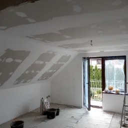 Pokój na poddaszu w trakcie szpachlowania: widoczne worki z gładzią, wiadra i zaklejona podłoga. Okno balkonowe z widokiem na zieleń.