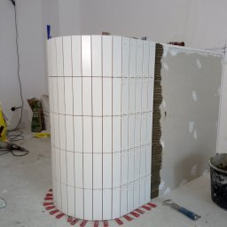 Piotr remonty - Zaokrąglona ściana wyłożona białymi, wąskimi płytkami w trakcie montażu. Widoczne czerwone kliny poziomujące i zaprawa. Prace glazurnicze w surowym wnętrzu.