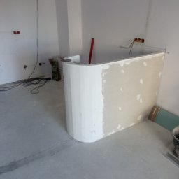 Piotr remonty - Narożnik z płyty gipsowej, częściowo wykończony gładzią, w trakcie prac wykończeniowych. Widoczne przewody elektryczne i torba z zaprawą.