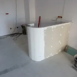 Narożnik z płyty gipsowej, częściowo wykończony gładzią, w trakcie prac wykończeniowych. Widoczne przewody elektryczne i torba z zaprawą.