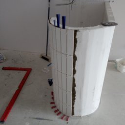 Piotr remonty - Montaż białych, pionowych płytek na zaokrąglonej konstrukcji w łazience. Widoczne narzędzia i materiały budowlane. W tle ściana z instalacją elektryczną.