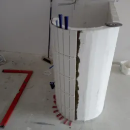 Montaż białych, pionowych płytek na zaokrąglonej konstrukcji w łazience. Widoczne narzędzia i materiały budowlane. W tle ściana z instalacją elektryczną.