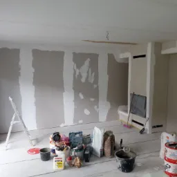 Wnętrze w trakcie prac wykończeniowych: ściany z częściowo nałożoną gładzią, narzędzia i materiały budowlane rozłożone na podłodze. Widoczna drabina i fragment instalacji kominkowej.