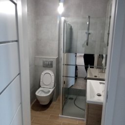 Piotr remonty - Nowoczesna łazienka w odcieniach szarości i beżu, z kabiną prysznicową ze szkła, podwieszaną toaletą i umywalką z szafką. Minimalistyczny design, brązowe płytki podłogowe.