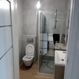 Nowoczesna łazienka w odcieniach szarości i beżu, z kabiną prysznicową ze szkła, podwieszaną toaletą i umywalką z szafką. Minimalistyczny design, brązowe płytki podłogowe.