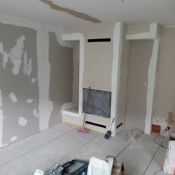 Piotr remonty - Wnętrze w trakcie szpachlowania: ściany z płyt gips-karton, fragment obudowy kominka, narzędzia i worki z zaprawą na podłodze. Widoczne niedoskonałości i etapy prac.