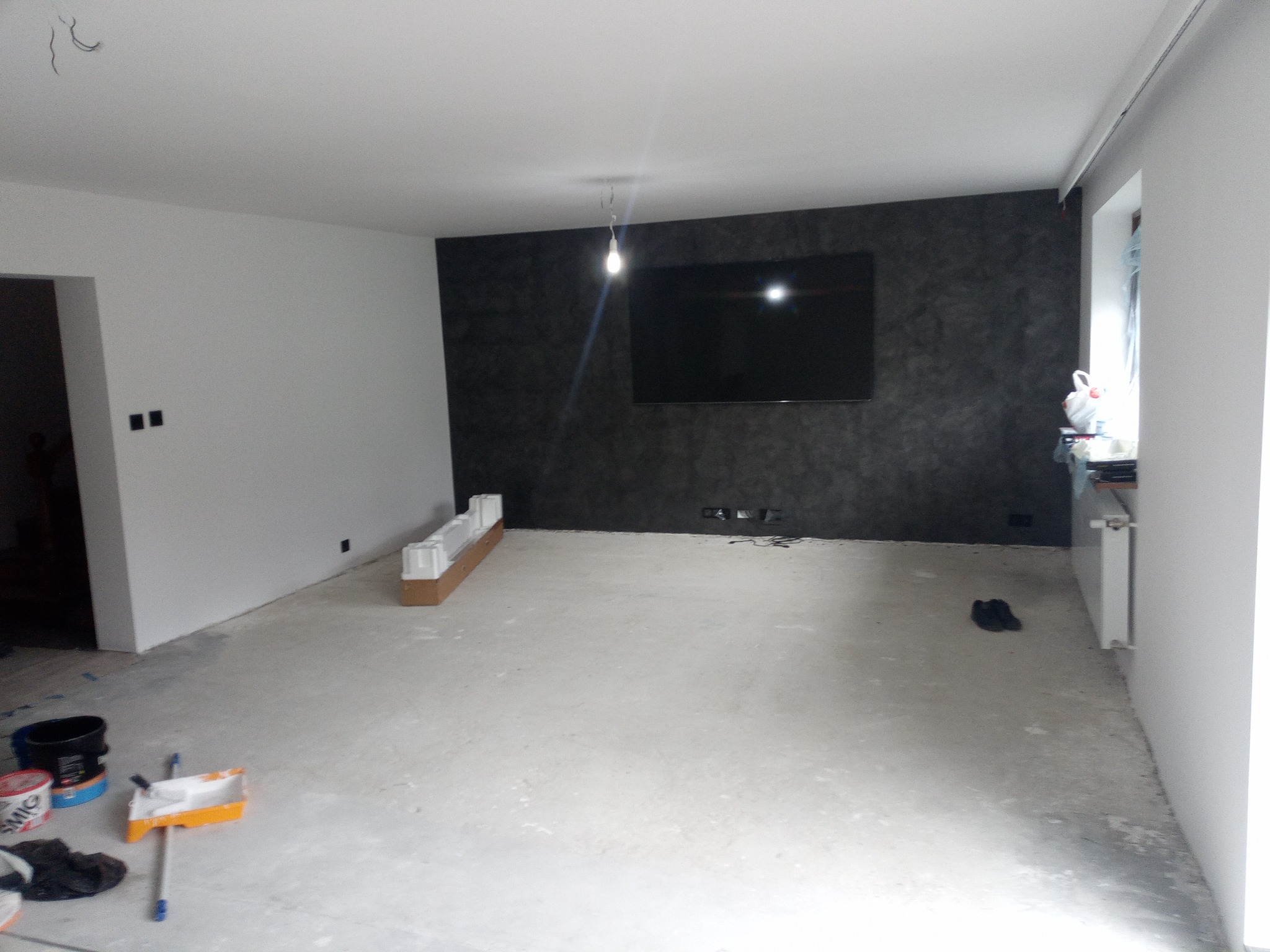 Pokój po remoncie: szare ściany, czarna ściana TV, narzędzia malarskie. Widoczny grzejnik i okno. Podłoga surowa, przygotowana do dalszych prac wykończeniowych.