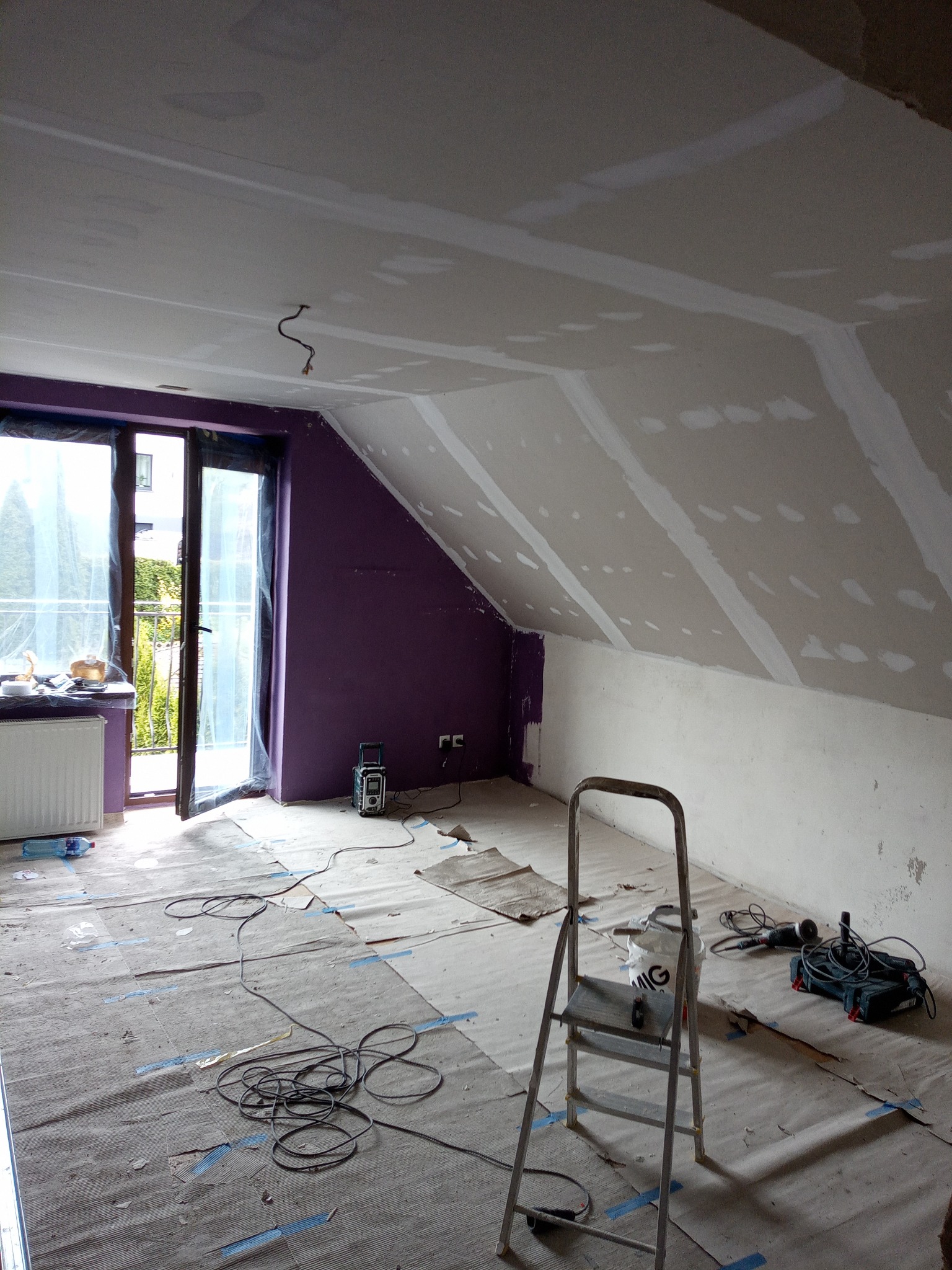 Poddasze w trakcie prac wykończeniowych: ściany i skosy pokryte płytami gips-karton, widoczne łączenia szpachlowane. Na podłodze rozłożona folia ochronna, drabina i narzędzia.