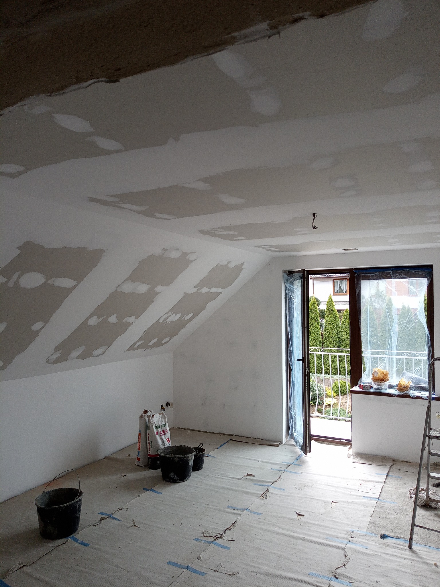 Pokój na poddaszu w trakcie szpachlowania: widoczne worki z gładzią, wiadra i zaklejona podłoga. Okno balkonowe z widokiem na zieleń.