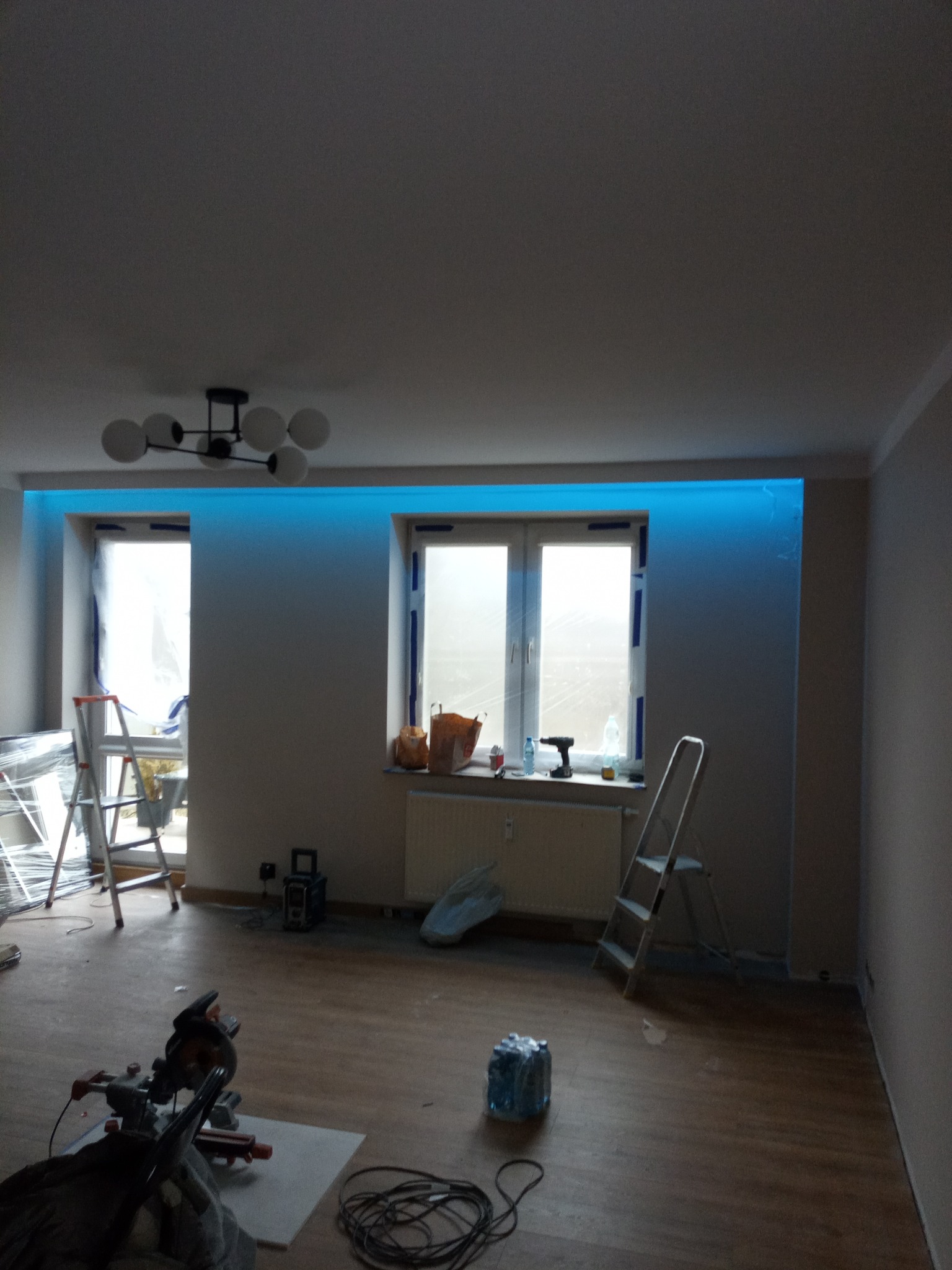 Wnętrze pokoju w trakcie prac wykończeniowych: widoczne drabiny, narzędzia, podświetlenie LED przy suficie, surowe ściany i podłoga. Etap przed finalnym umeblowaniem.