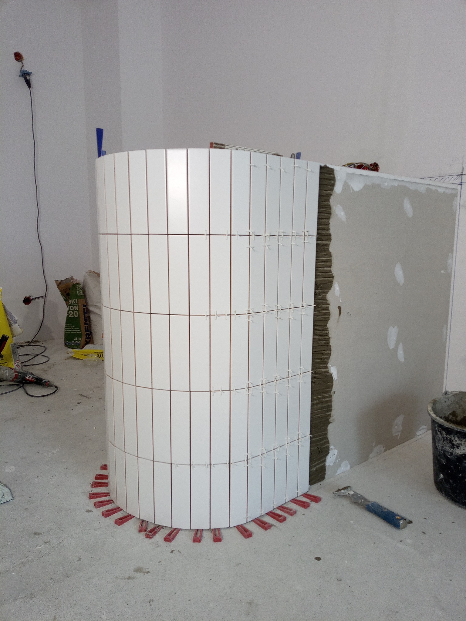 Zaokrąglona ściana wyłożona białymi, wąskimi płytkami w trakcie montażu. Widoczne czerwone kliny poziomujące i zaprawa. Prace glazurnicze w surowym wnętrzu.