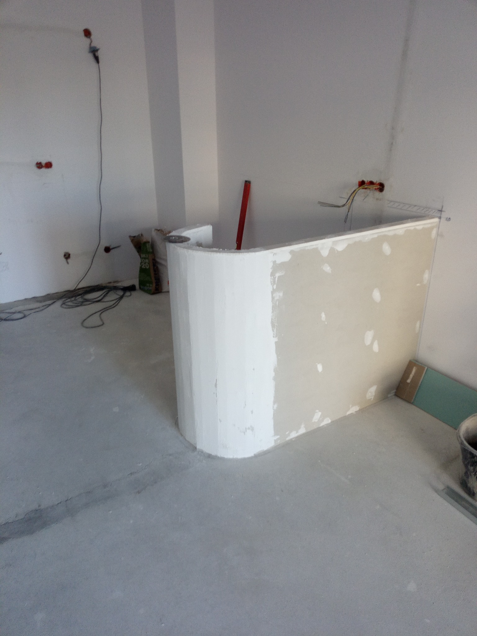 Narożnik z płyty gipsowej, częściowo wykończony gładzią, w trakcie prac wykończeniowych. Widoczne przewody elektryczne i torba z zaprawą.