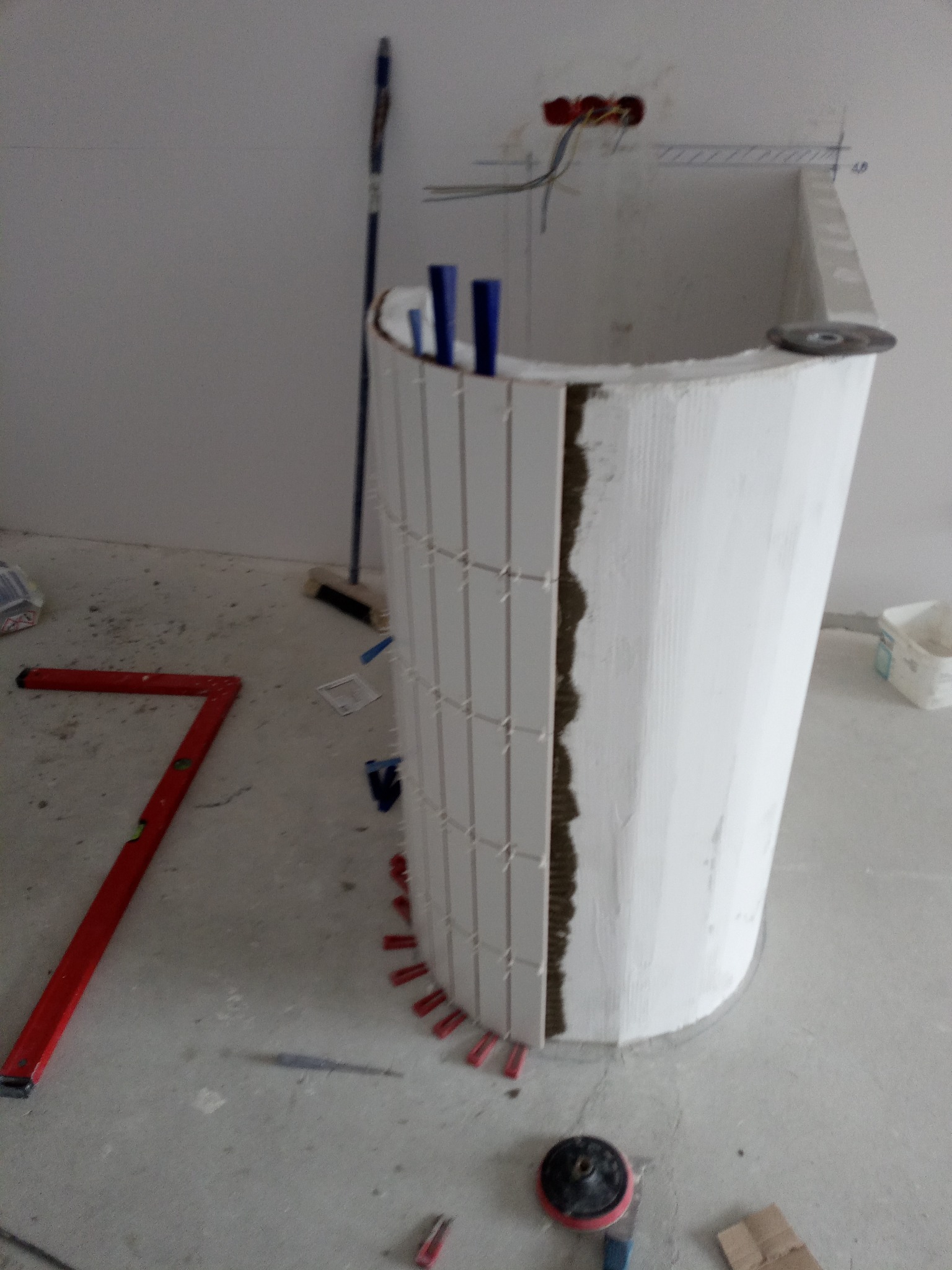 Montaż białych, pionowych płytek na zaokrąglonej konstrukcji w łazience. Widoczne narzędzia i materiały budowlane. W tle ściana z instalacją elektryczną.