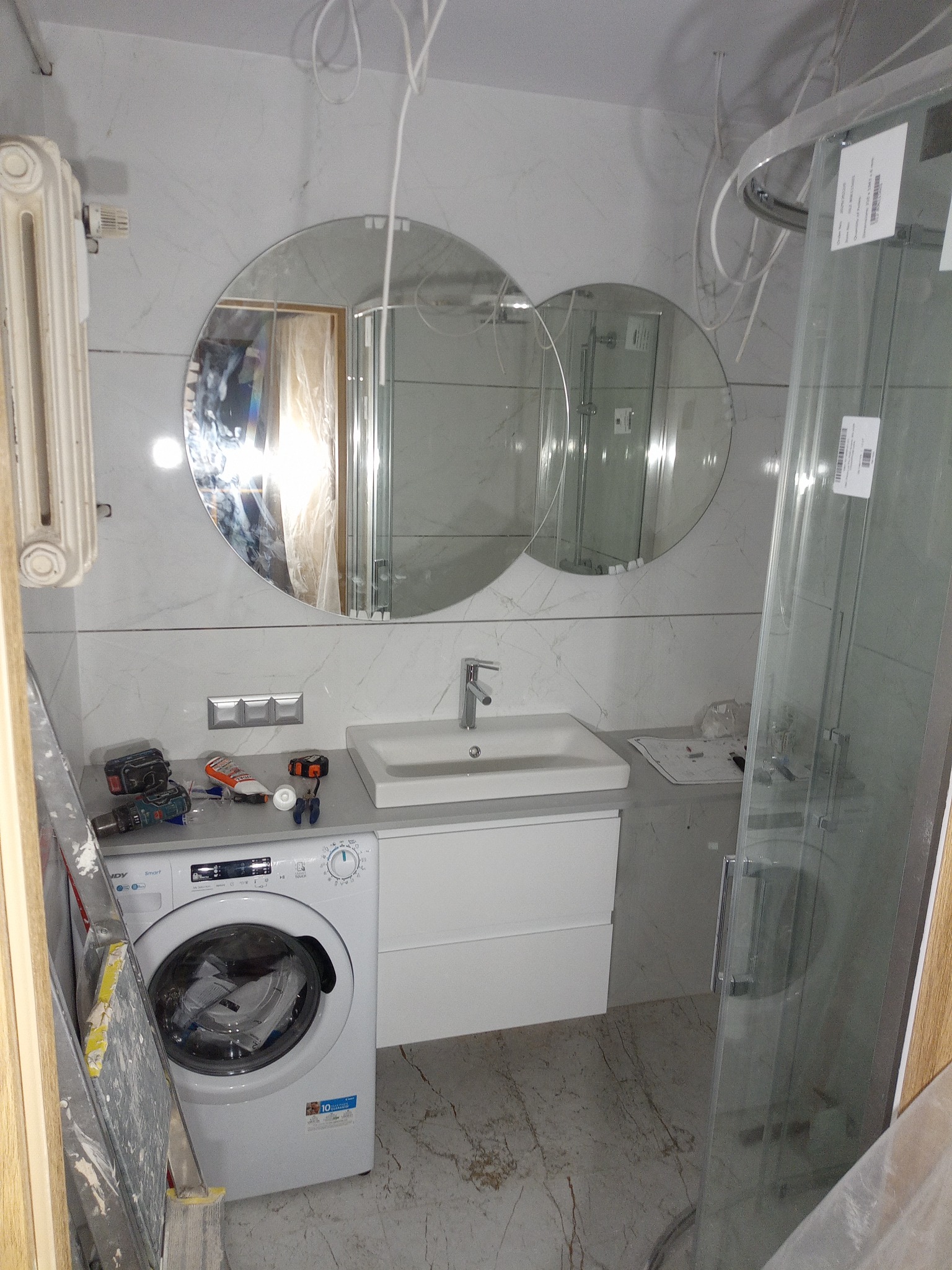 Remont łazienki w Słupsku: widok na umywalkę z szafką, okrągłe lustra, kabinę prysznicową i pralkę. Widoczne narzędzia i drabina, co sugeruje trwający remont.
