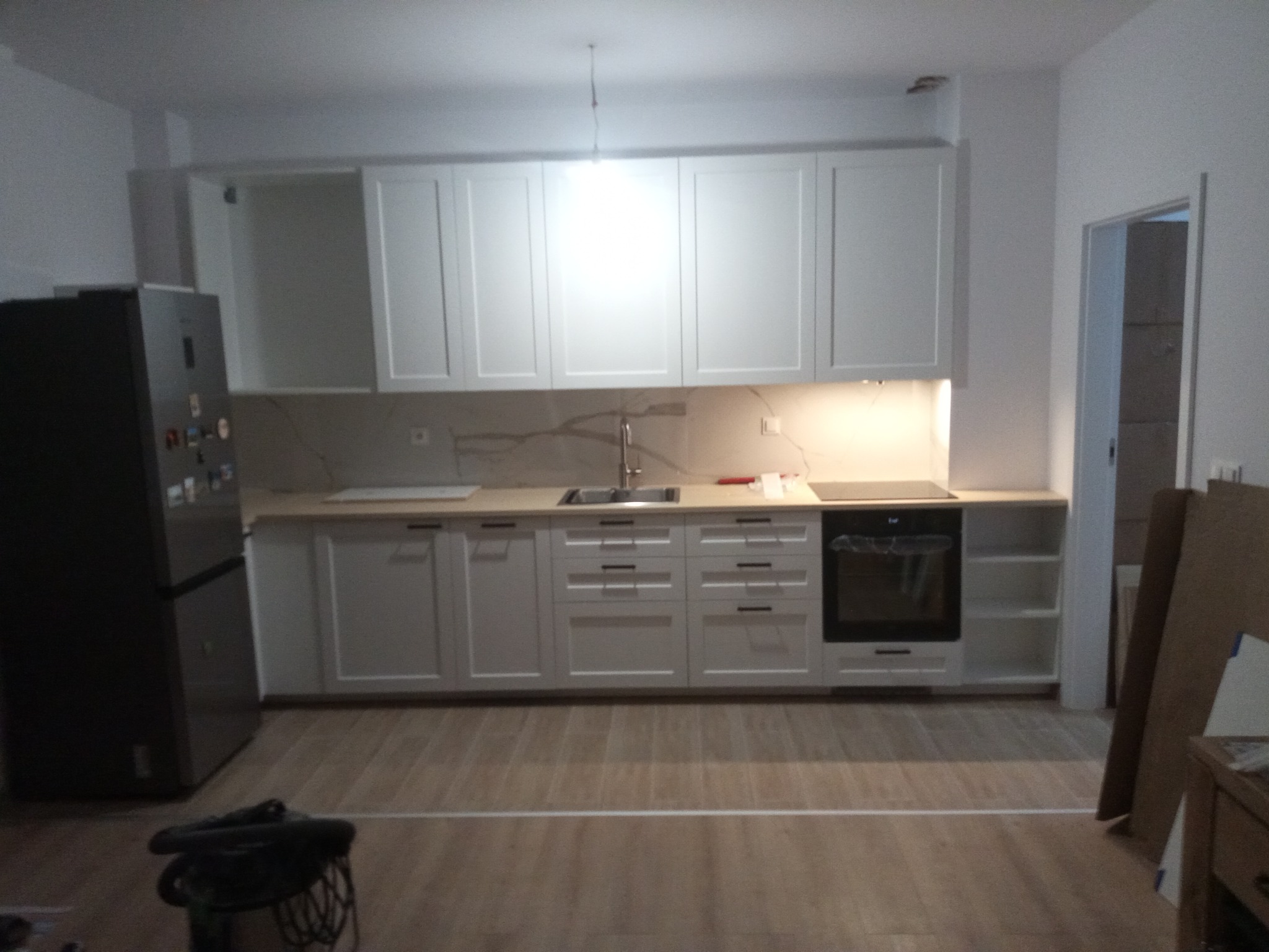 Jasna kuchnia z drewnianą podłogą, białymi szafkami i sprzętem AGD. Widoczny czarny, duży lodówka, jasny blat i oświetlenie LED pod szafkami.