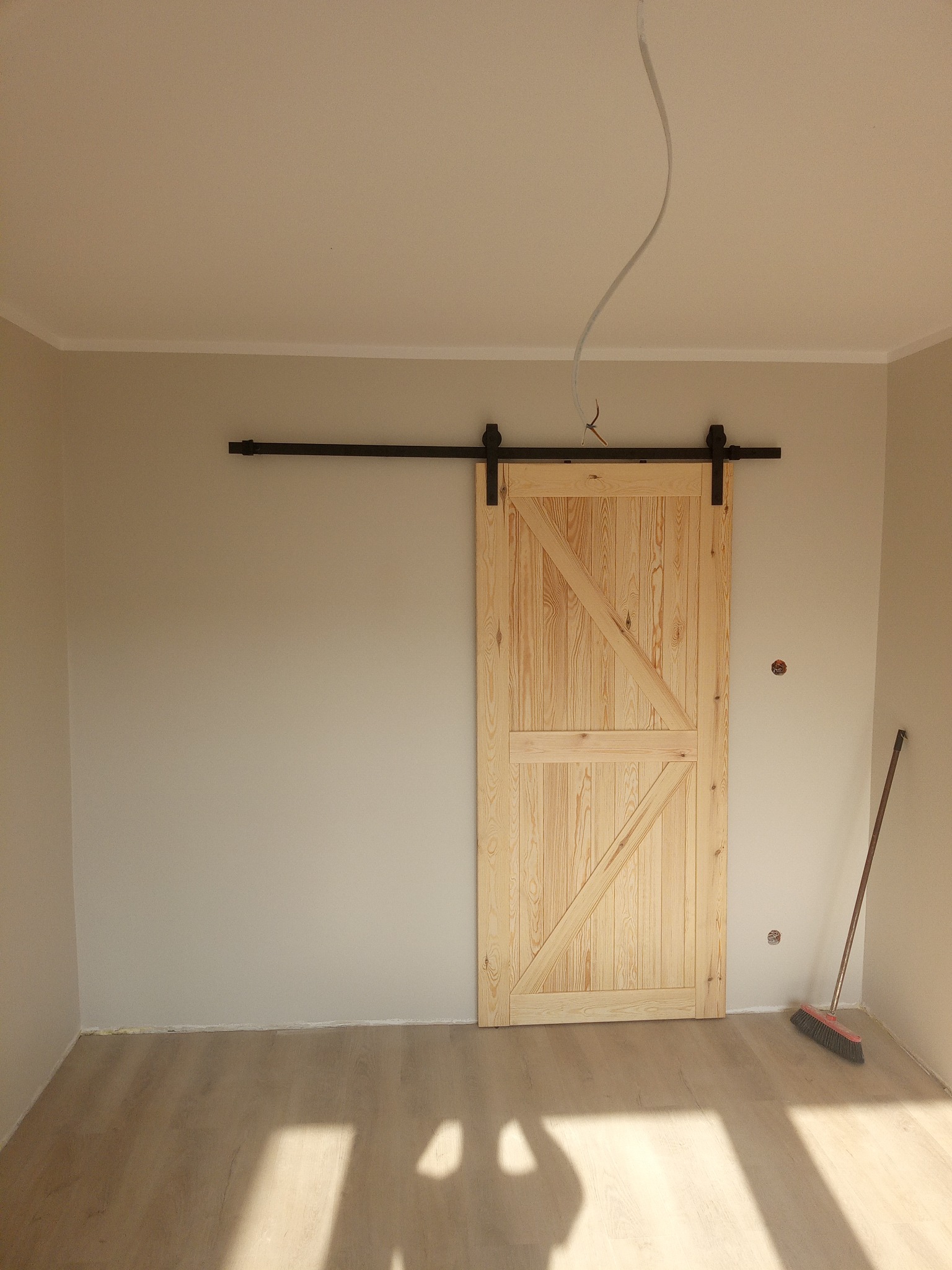 Jasne, drewniane drzwi przesuwne w stylu rustykalnym na czarnej szynie, zamontowane w pokoju z panelami i beżowymi ścianami. Widoczny wystający kabel i miotła.