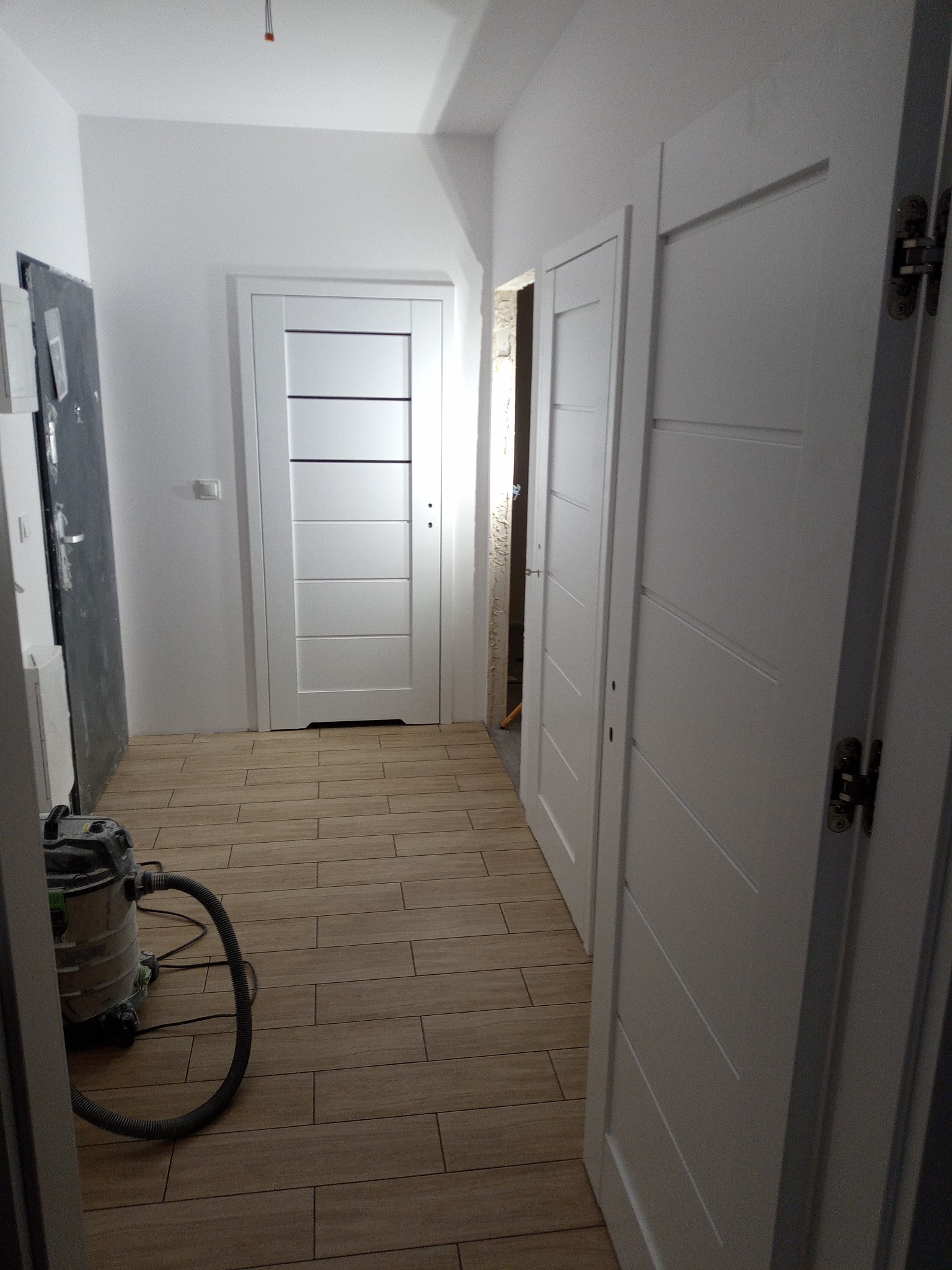 Jasne wnętrze po remoncie: drewnopodobna podłoga, białe drzwi z poziomymi frezami, odkurzacz budowlany. Widok z perspektywy wejścia do pomieszczenia.