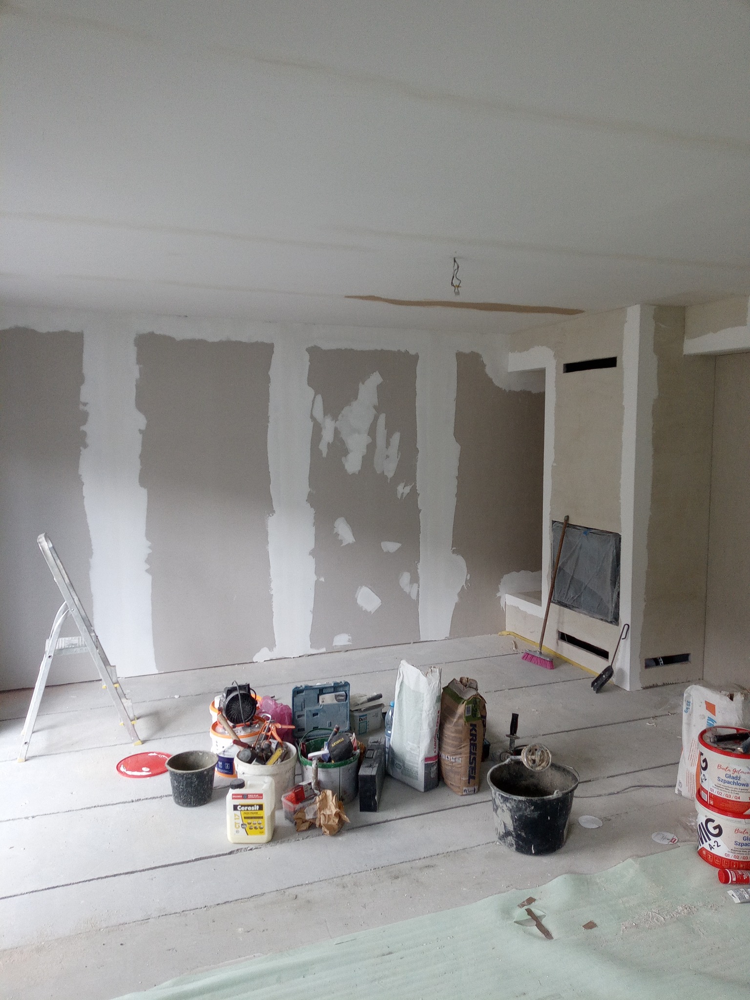 Wnętrze w trakcie prac wykończeniowych: ściany z częściowo nałożoną gładzią, narzędzia i materiały budowlane rozłożone na podłodze. Widoczna drabina i fragment instalacji kominkowej.