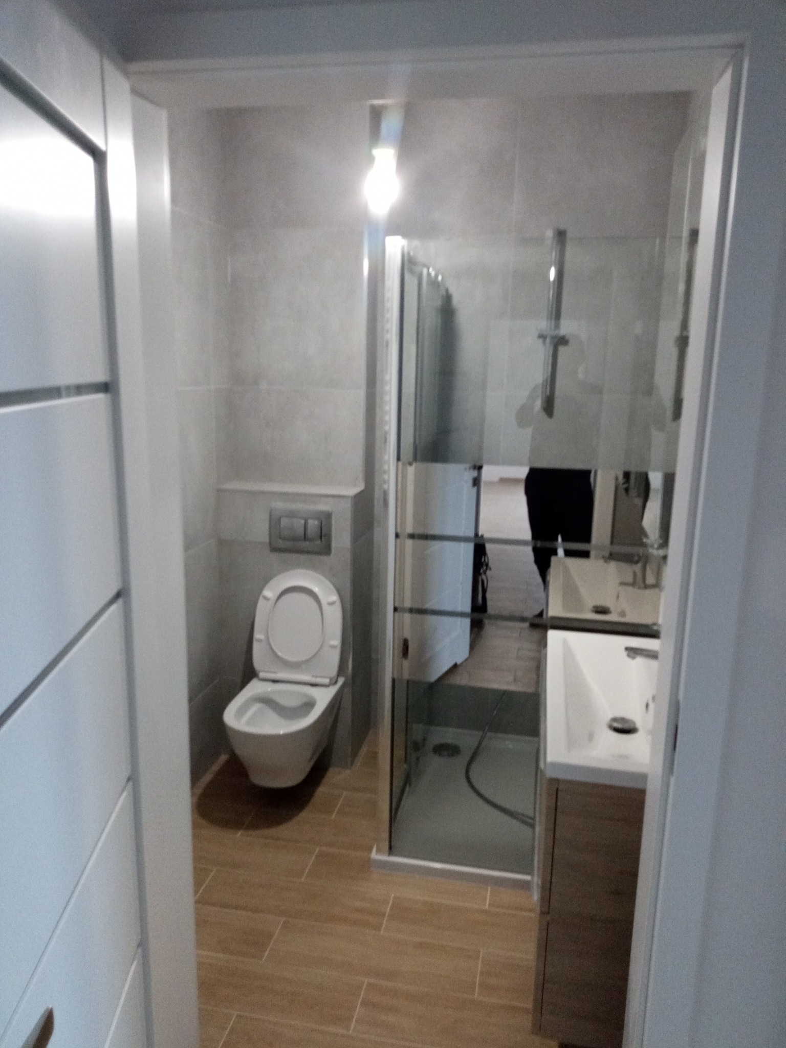 Nowoczesna łazienka w odcieniach szarości i beżu, z kabiną prysznicową ze szkła, podwieszaną toaletą i umywalką z szafką. Minimalistyczny design, brązowe płytki podłogowe.