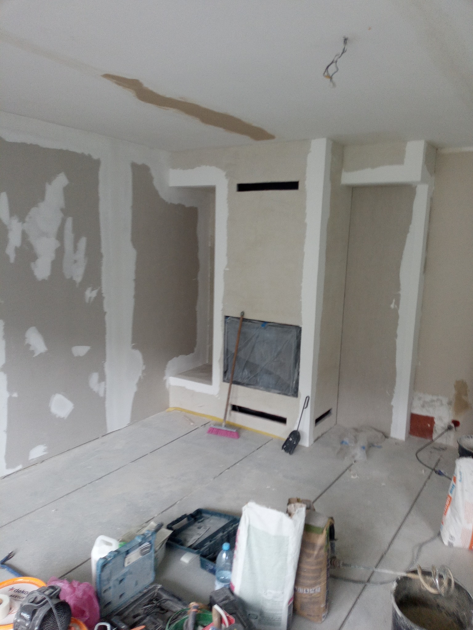 Wnętrze w trakcie szpachlowania: ściany z płyt gips-karton, fragment obudowy kominka, narzędzia i worki z zaprawą na podłodze. Widoczne niedoskonałości i etapy prac.