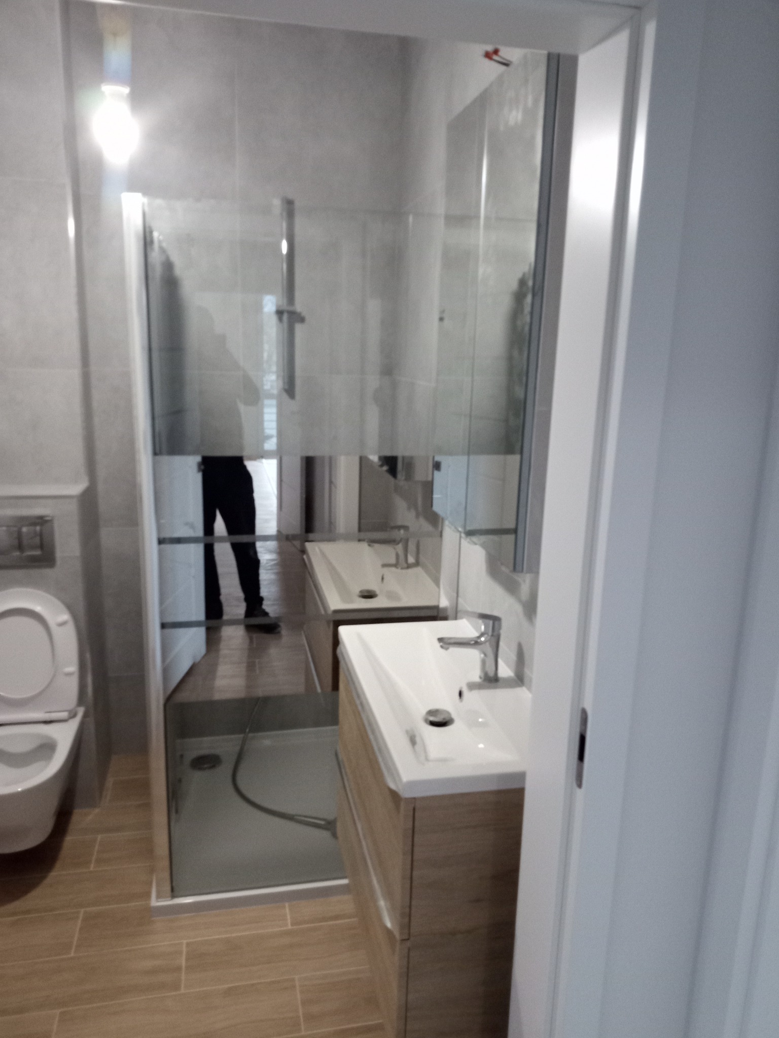 Nowoczesna, jasna łazienka po remoncie: kabina prysznicowa ze szklaną ścianką, umywalka z szafką w kolorze drewna i toaleta. Widoczne płytki podłogowe imitujące drewno.