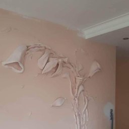 Zibination. Art - Niedokończona dekoracja ścienna z gładzi gipsowej w kształcie kwiatów lilii, widoczne ślady szpachlowania, blade kolory, fragment listwy sufitowej.