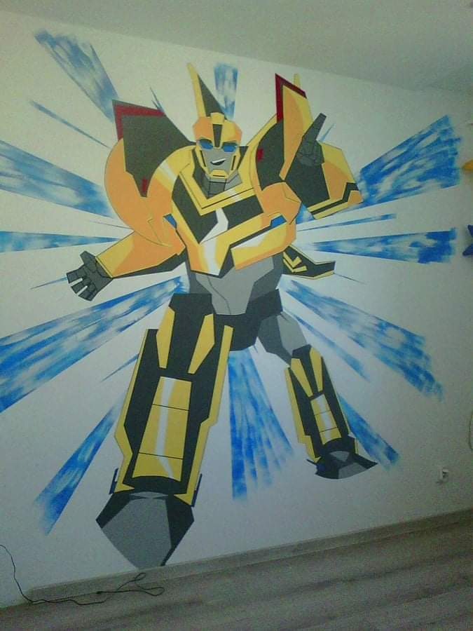 Malowidło ścienne przedstawiające postać Bumblebee z Transformers w pokoju dziecięcym, wykonane farbami w odcieniach żółci, szarości i błękitu, z efektem rozchodzących się promieni.