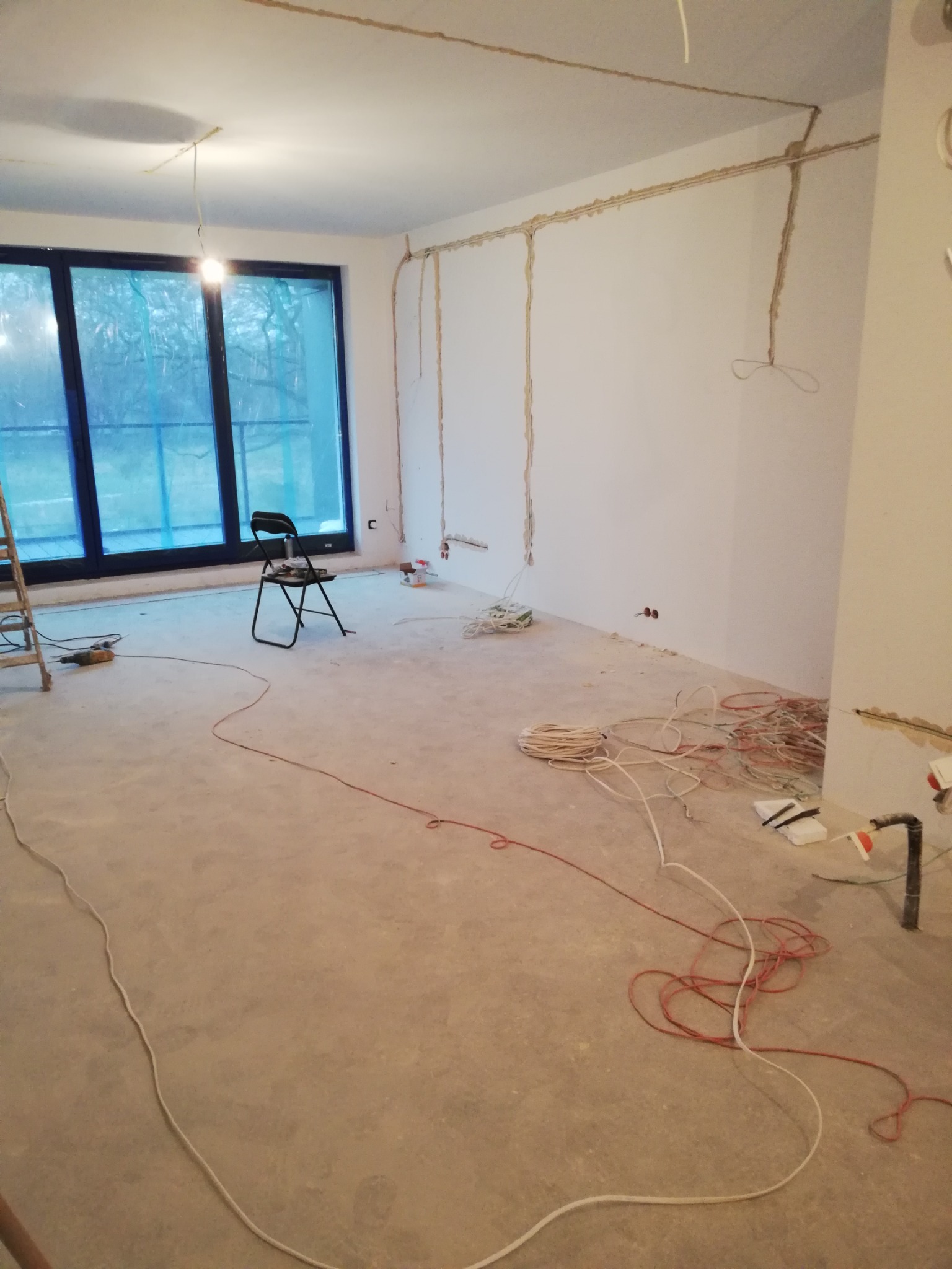 Surowy stan wnętrza w trakcie prac instalacyjnych: wyżłobione bruzdy w ścianach pod przewody elektryczne, zwisające kable, rozłożone przewody na podłodze, drabina oparta o okno, krzesło na środku...